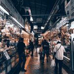 Consumidores circulam por um corredor movimentado de um mercado coberto, onde há diversas lojas e bancas.