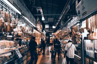 Consumidores circulam por um corredor movimentado de um mercado coberto, onde há diversas lojas e bancas.