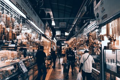 Consumidores circulam por um corredor movimentado de um mercado coberto, onde há diversas lojas e bancas.
