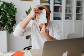 Empresária mostra um recibo em um ambiente de trabalho doméstico, com computador e documentos ao redor.