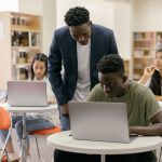Estudantes de diferentes perfis participam de uma aula enquanto uma professora os orienta, com notebooks sobre as mesas.