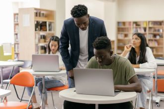 Estudantes de diferentes perfis participam de uma aula enquanto uma professora os orienta, com notebooks sobre as mesas.